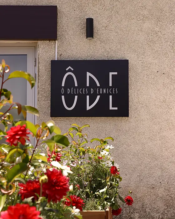 L'enseigne d'un commerce sur un mur crépi, dahlias rouges en contrebas